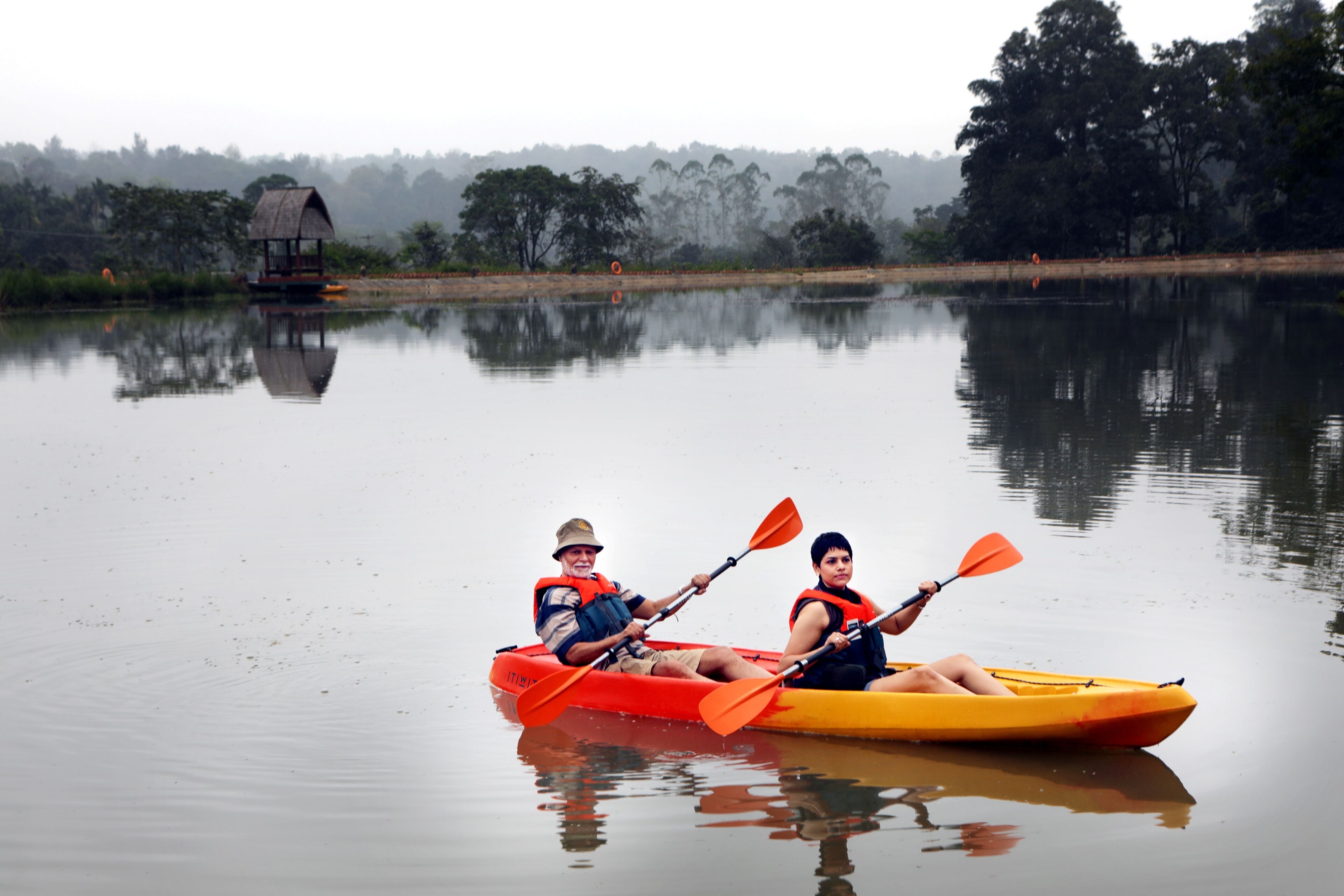 Two people kayaking on a calm lake with a hut in the background.