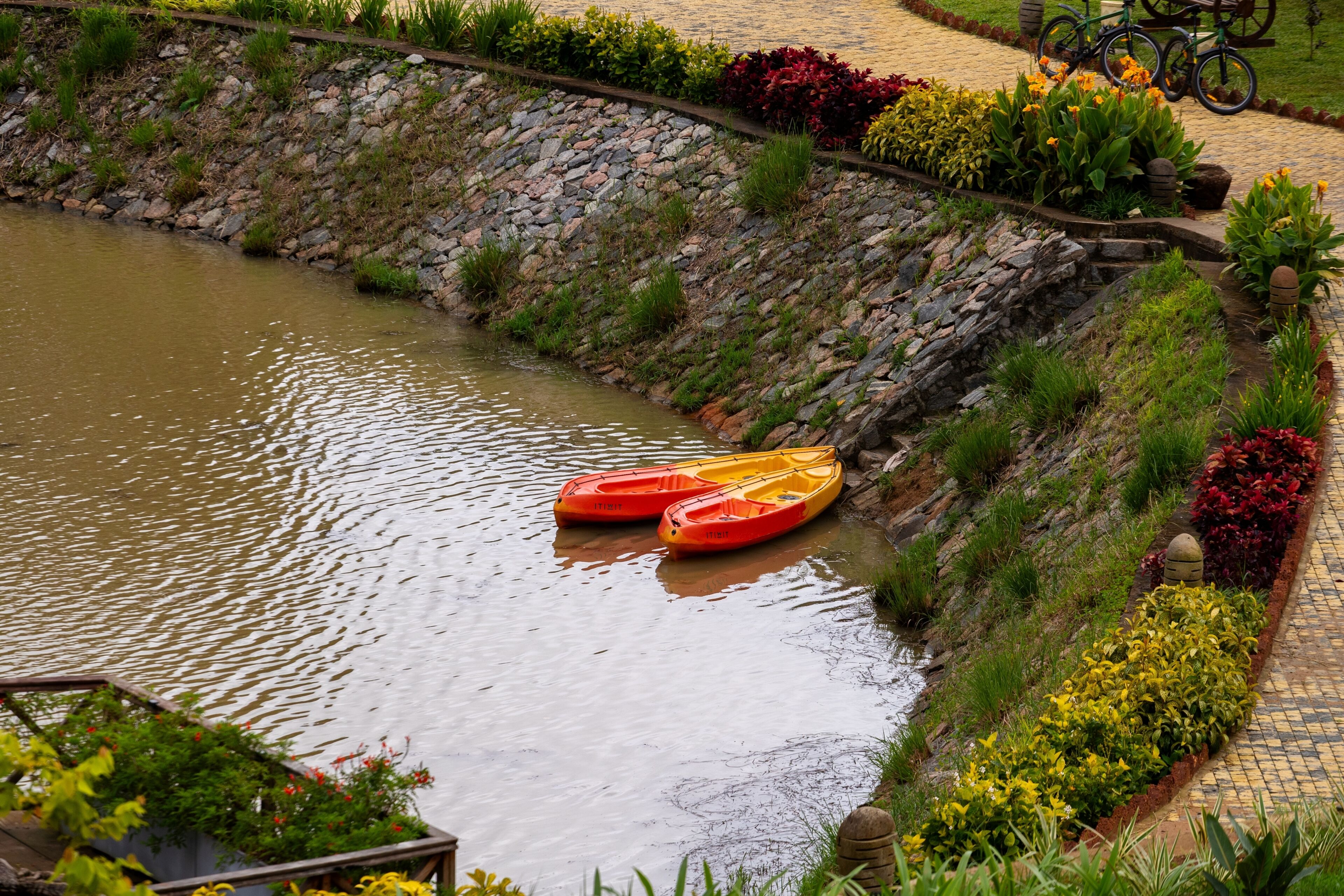 A river with two kayaks, a stone wall, and a pathway with plants.