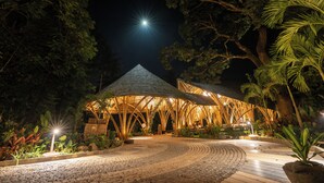 A night-time outdoor pavilion with a wooden roof structure, surrounded by tropical plants and a cobblestone pathway. - The Leela Coorg Forest Sanctuary (Horur)