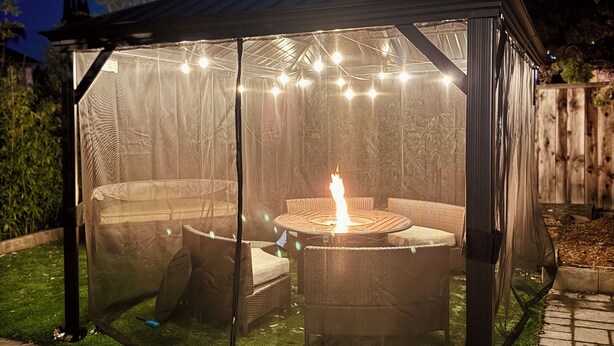 A covered outdoor seating area with a fire feature, surrounded by a mesh enclosure and string lights. - Modern 2BR Near SJC | W/D | FirePit | Free Parking (San Jose)
