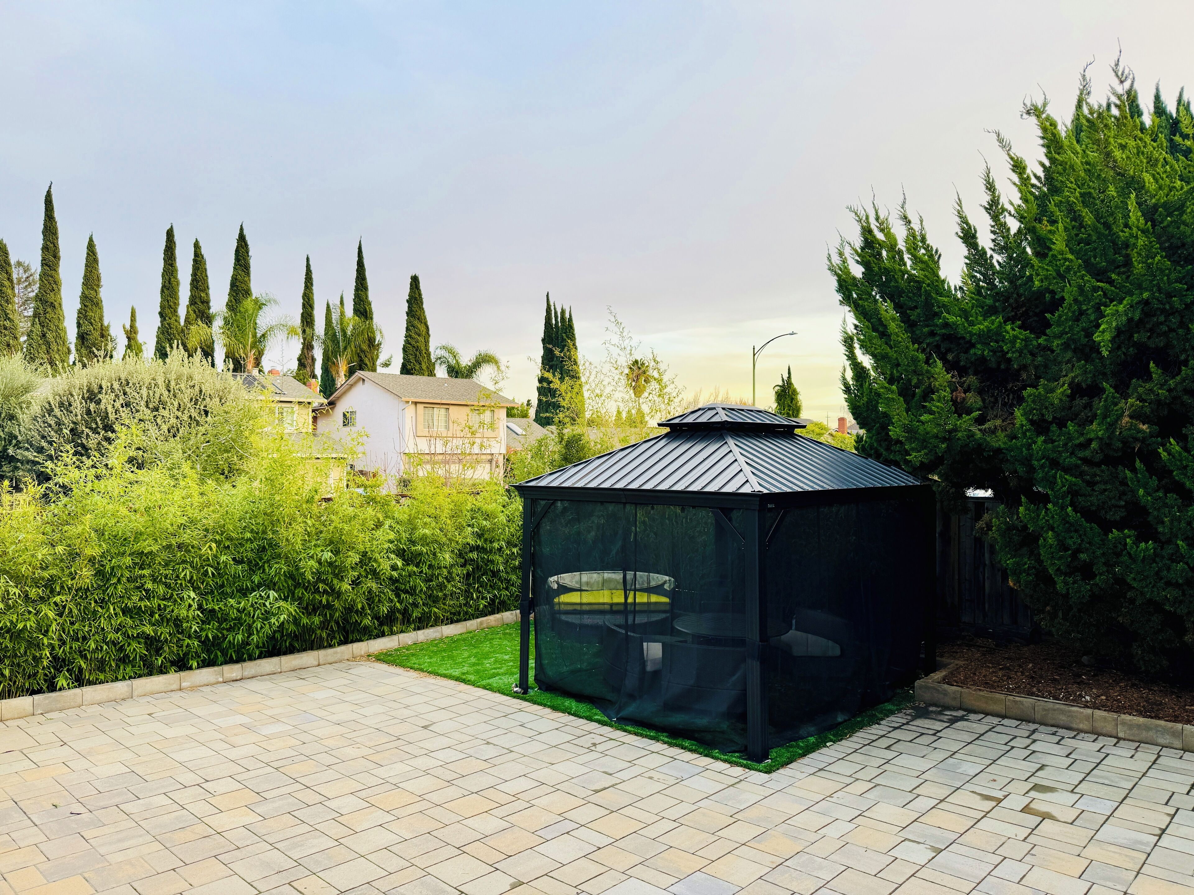 Um gazebo com telhado de metal e paredes de vidro, rodeado por um pátio de tijolos e vegetação exuberante.