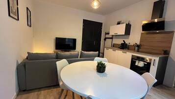 Un ambiente compatto con angolo pranzo, divano, televisore, angolo cottura e lampada a sospensione.