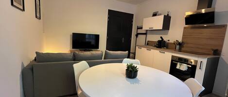A compact living space with a dining area, a sofa, a TV, a kitchenette, and a hanging light fixture.