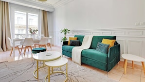 Uma sala de estar moderna com um sofá verde, detalhes em branco e dourado, um espaço de refeições e uma grande janela com cortinas.