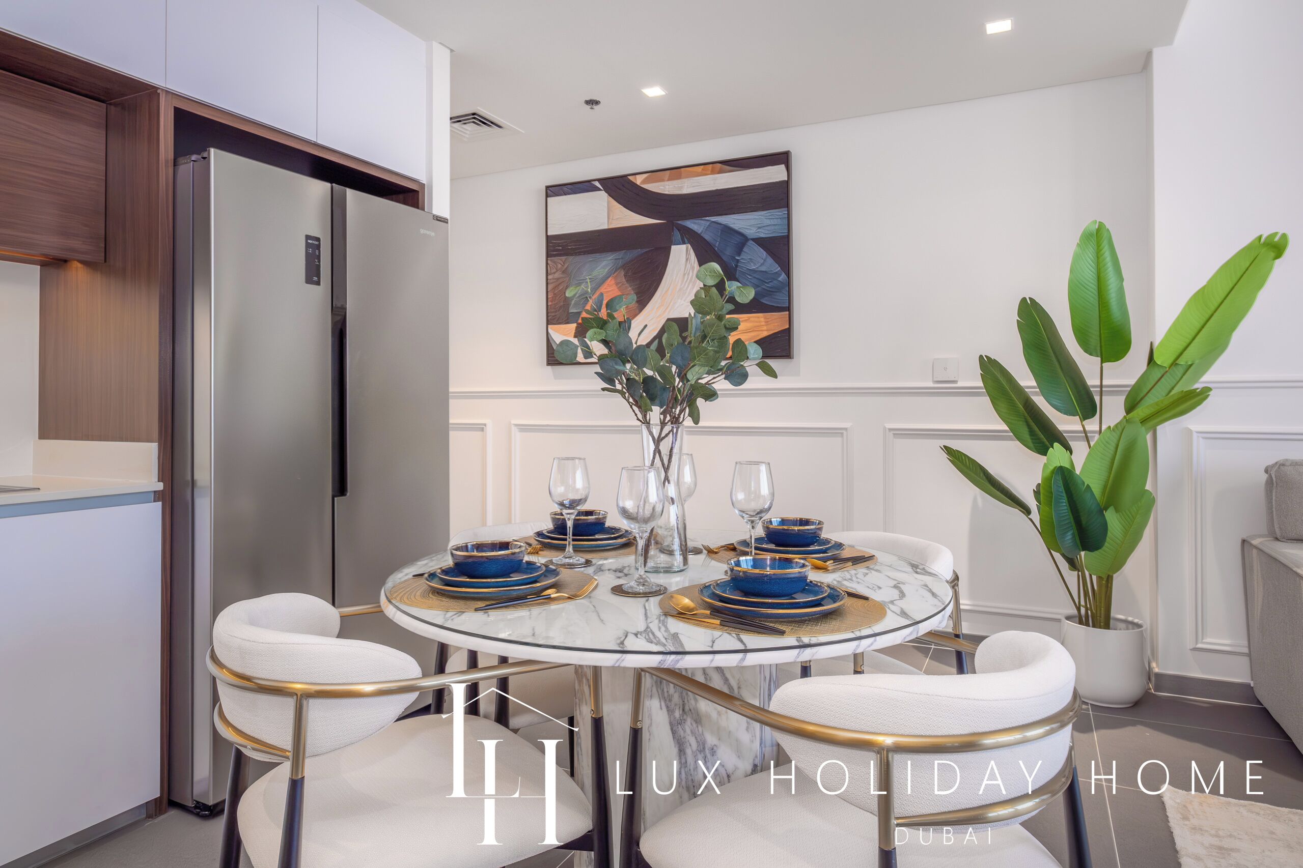 A modern dining area with a round table set for four, featuring blue and gold dinnerware, white upholstered chairs, and a marble top.