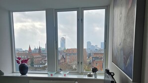A bedroom with a bed, a desk, a chair, a radiator, a window with a view of buildings, and a painting on the wall. - Central apartment with panoramic view and roof terrace   (Aarhus)
