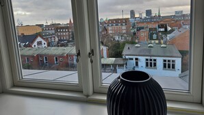 A view through a window with a black vase on the sill, overlooking a cityscape with various buildings and a church spire. - Central apartment with panoramic view and roof terrace   (Aarhus)