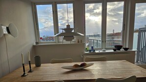 A dining area with a wooden table, two chairs, a white pendant light, and a view of the city through large windows. - Central apartment with panoramic view and roof terrace   (Aarhus)