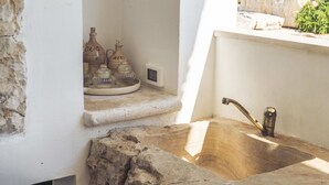 A stone sink with a metal faucet, a small shelf with glass jars labeled "OLIO" and "ACETO," and a power outlet on the wall.