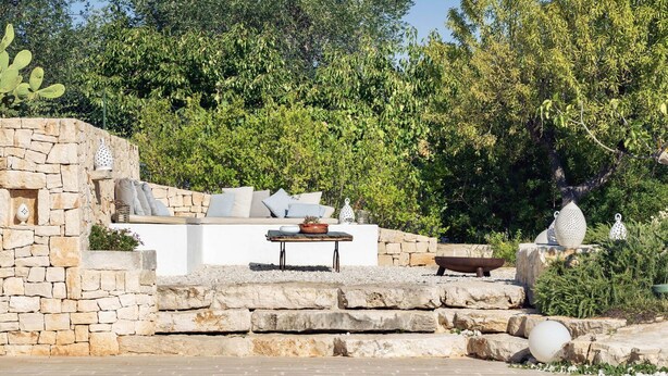 A stone-built outdoor seating area with a table and cushions, overlooking a pool.