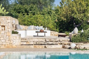 Un espace extérieur aménagé avec des sièges en pierre, une table et des coussins, donnant sur une piscine.