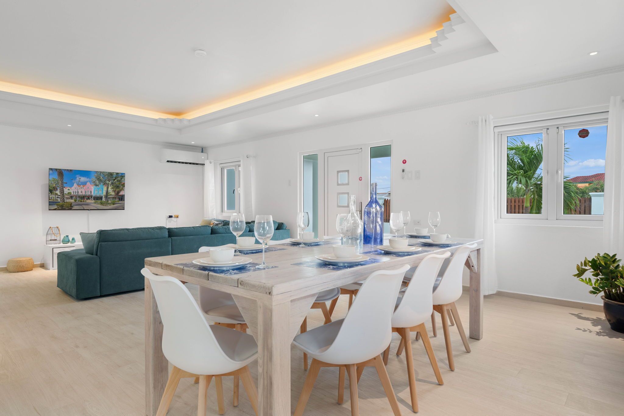 Un comedor moderno con una mesa de madera dispuesta para seis personas, sillas blancas y un sofá color turquesa al fondo.