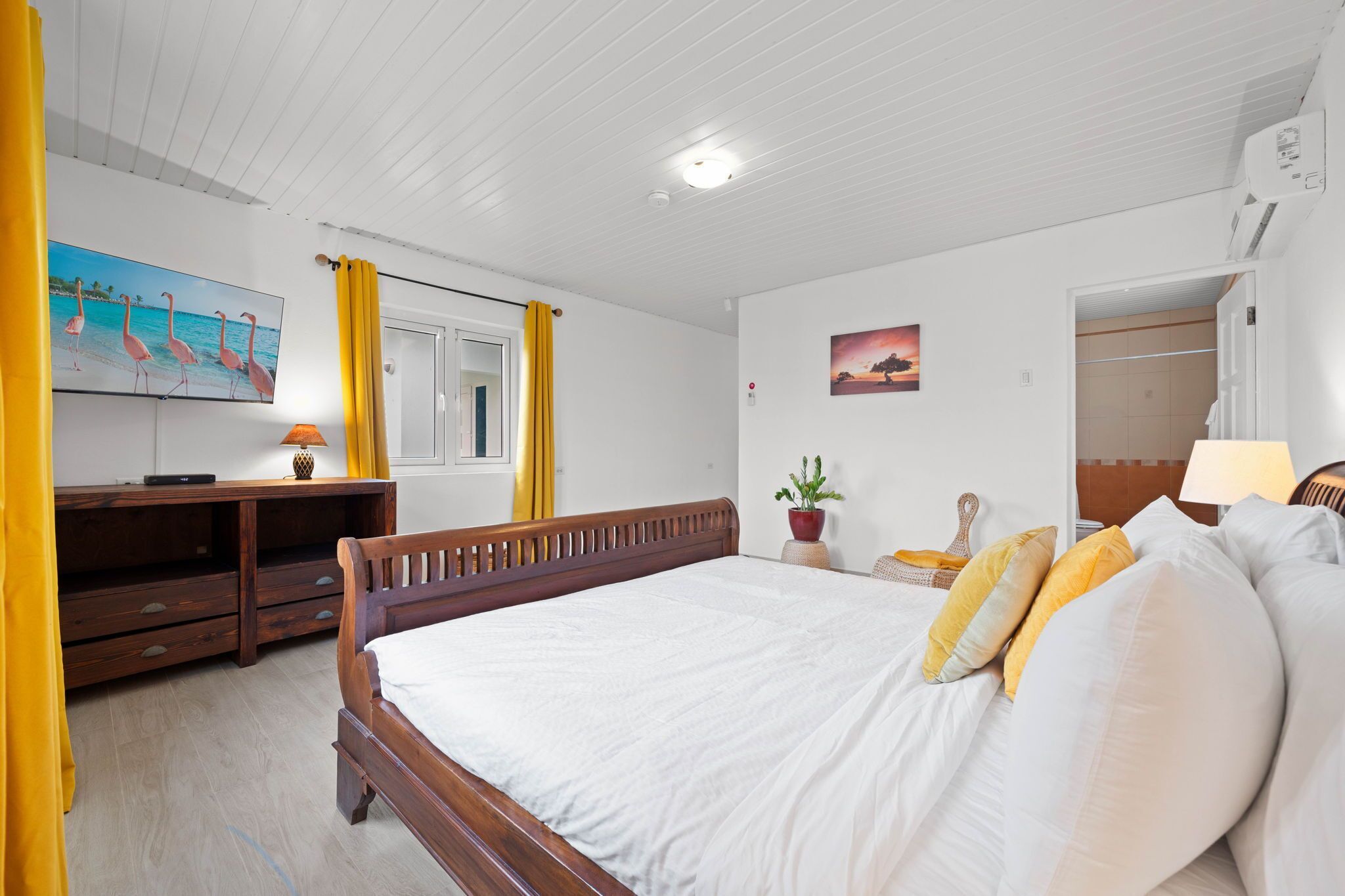 Een slaapkamer met een houten bed, een televisie op een houten standaard, een kamerplant en een schilderij aan de muur.