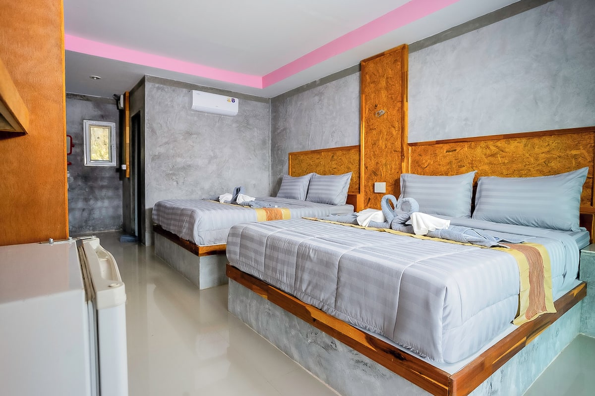 A hotel room with two beds, a wooden headboard, a white refrigerator, and a grey wall with an air conditioning unit.