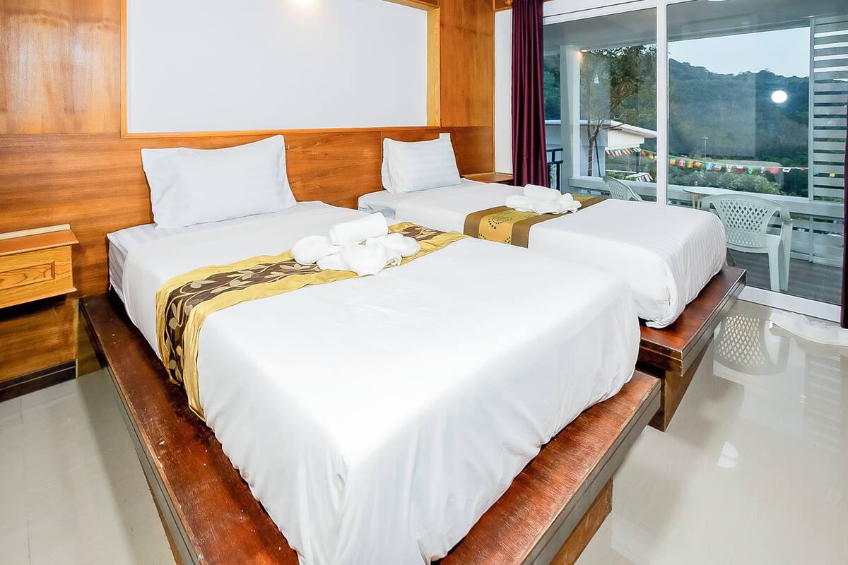 A hotel room with two beds, wooden headboard, large windows, and a balcony with a view of the landscape.
