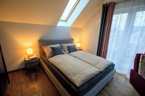 Un dormitorio con tragaluz, una cama con dos almohadas, una mesita de noche con lámpara y una ventana con cortinas.