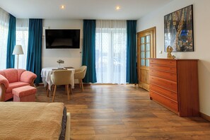 A hotel room with a bed, a wooden dresser, a TV mounted on the wall, a dining table with chairs, and a window with curtains.