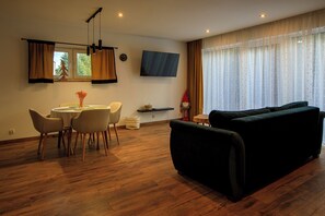 Uma sala de estar moderna com um sofá escuro, uma área de jantar com uma mesa redonda e cadeiras, e uma TV fixada na parede.