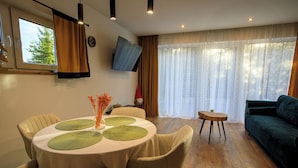 A dining area with a round table, chairs, and a vase of flowers. There is a sofa and a TV mounted on the wall.