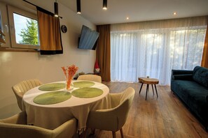 A dining area with a round table, chairs, and a vase of flowers. There is a sofa and a TV mounted on the wall.