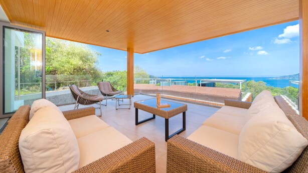 A modern outdoor lounge area with wicker furniture and a glass coffee table, overlooking a scenic view of the sea. - Luxury 2BR Apartment in Bluepoint 82 (Patong)
