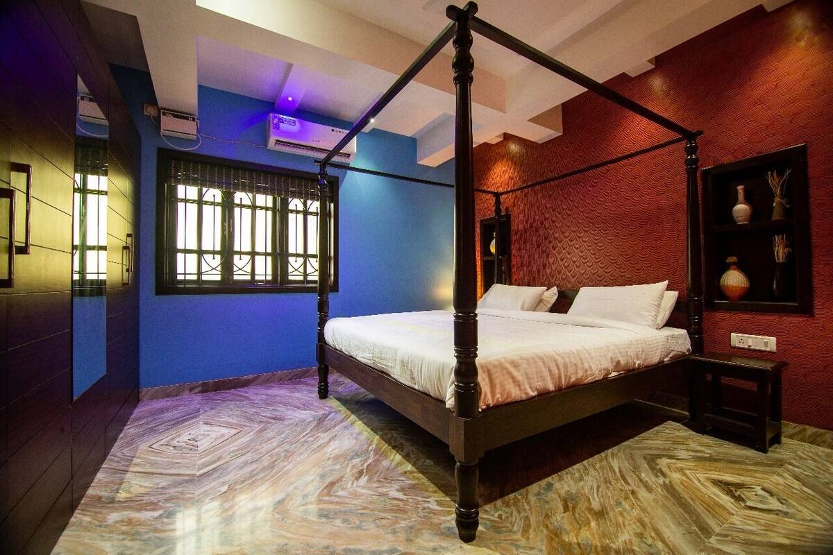 Una cama con dosel en una habitación con paredes azules y rojas, suelo estampado y una ventana con rejas.