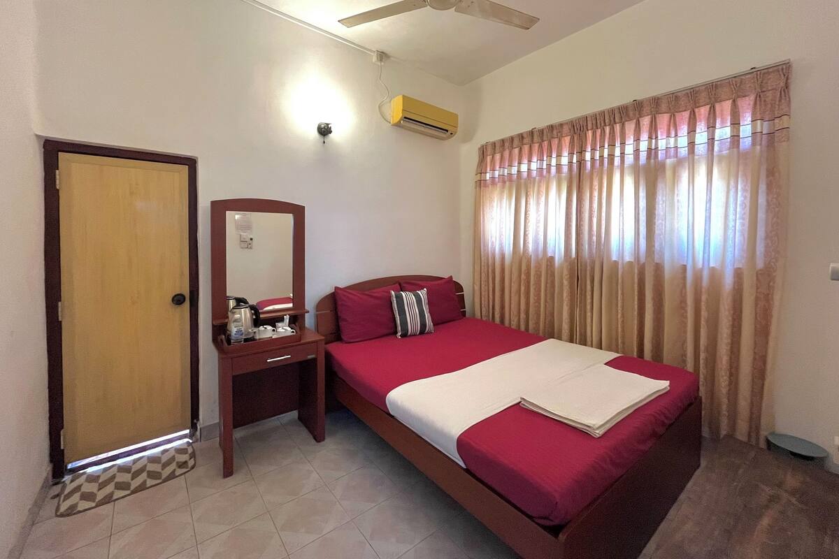 Habitación de hotel con cama, escritorio, espejo, ventilador de techo y ventana con cortinas.