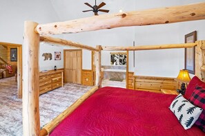 A room with a wooden ceiling structure, a bed with a red bedspread, a nightstand with a lamp, and a view of a forest through a window.