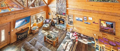 L’intérieur d’une cabane en rondins, avec une cheminée en pierre, des murs en bois, une grande fenêtre et un coin salon confortable.