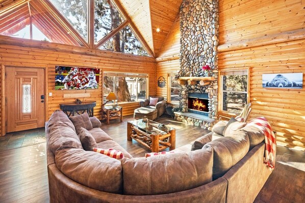 A log cabin interior with a curved leather sofa, a stone fireplace, and a television mounted on the wall.