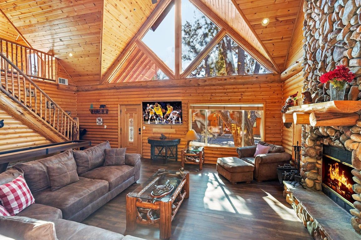 A log cabin interior with a wooden staircase, a fireplace, and a large window.