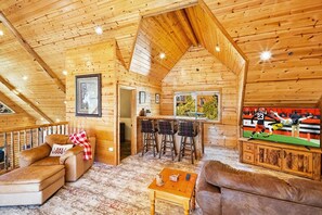 Interior acogedor de una cabaña con techo de madera, una zona de bar y un televisor mostrando un evento deportivo.