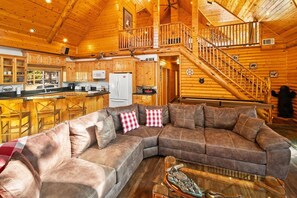 A spacious living area with a large sectional sofa, a kitchen with wooden cabinets, and a staircase leading to an upper level.