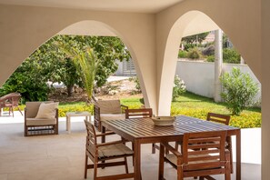 Un patio al aire libre con una mesa y sillas de madera, rodeado de arcos, y vistas a zonas verdes y un edificio lejano.