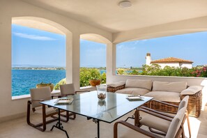 Una habitación iluminada por el sol con una mesa de vidrio, sillas, un sofá y vistas al mar y un edificio.