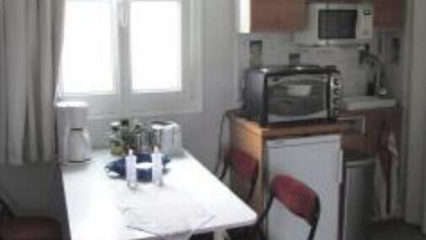 Una cuina compacta amb electrodomèstics bàsics, taula de dinar amb caires i finestra amb cortines.