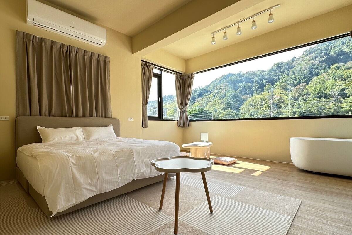 A bedroom with a bed, a round table, a bathtub, and a large window with a forest view.