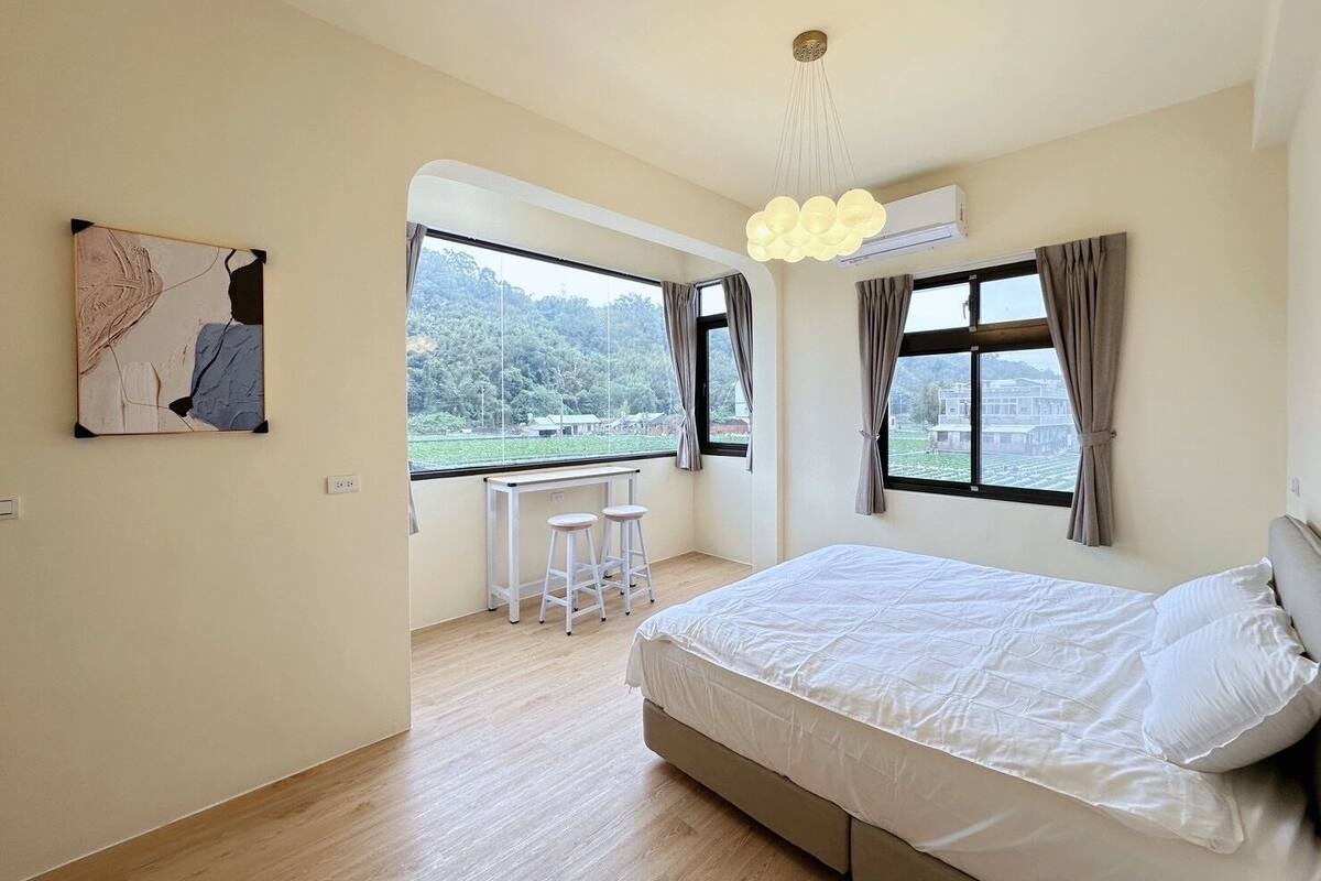 A bedroom with a bed, a desk with two stools, a large window with a view of a forest, and an air conditioning unit.