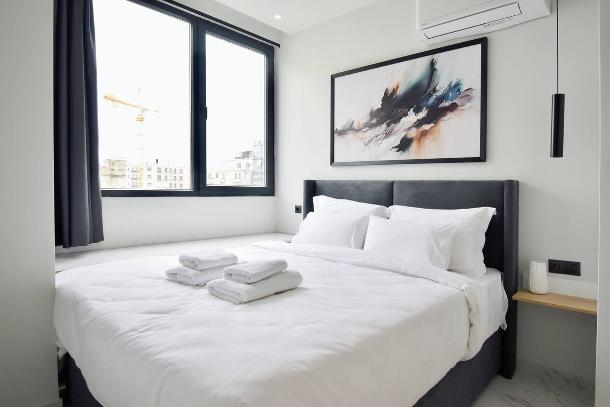 Ein modernes Hotelzimmer mit einem großen Bett, Blick auf die Stadt durch das Fenster und einem modernen, abstrakten Gemälde an der Wand.