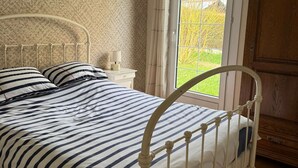A bedroom with a bed, a nightstand, a window with curtains, and a wooden wardrobe. - Le Rivage, House with garden near the sea (Saint-Aubin-sur-Mer)