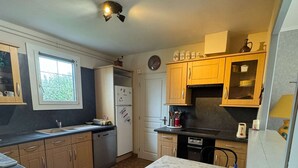 A compact kitchen with a dining table, hexagonal patterned floor, and wooden cabinets. - Le Rivage, House with garden near the sea (Saint-Aubin-sur-Mer)