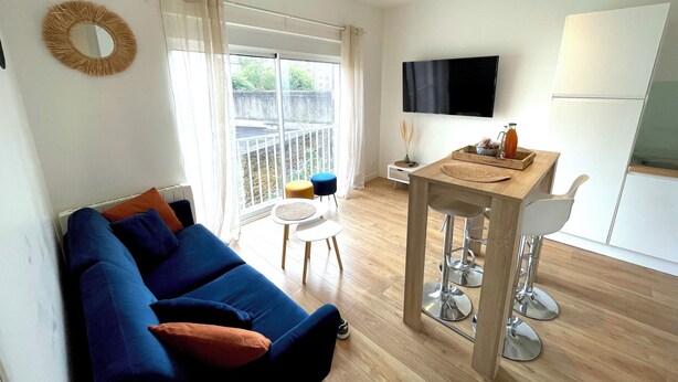 A modern living room with a blue sofa, a small dining table with two chairs, a TV mounted on the wall, and a balcony with a view. - Apartment "Le Clémenceau", downtown, fully equipped (Saint-Brieuc)