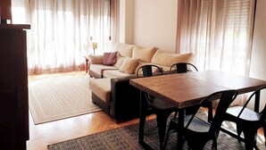 A cozy living room with a sofa, a dining table, and chairs. There are curtains, a lamp, and a ceiling light fixture. - Innhome - Jaime del Amo (Suances)