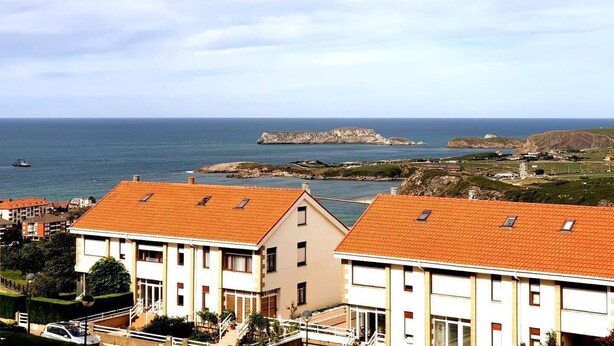 A coastal view with buildings featuring red-tiled roofs and a rocky outcrop in the distance. - Innhome - Jaime del Amo (Suances)