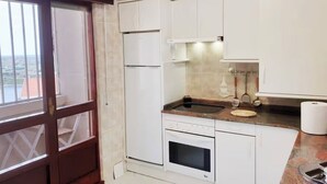 A compact kitchen with white cabinets, a built-in oven, a sink, and a window with a view of a red-roofed building. - Innhome - Jaime del Amo (Suances)