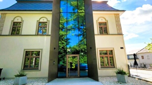 A building with a central glass facade, a traditional roof, and a symmetrical design with two visible windows on each side. - Apartment in villa, sky view, elevator, luxury floor (Bad Hall)