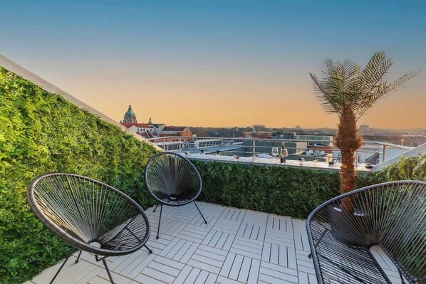 A rooftop terrace with two black wire chairs, a palm tree, and a view of a cityscape including a church.