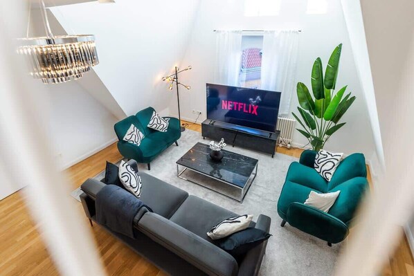A modern living room with a flat-screen TV, two teal armchairs, a dark sofa, a coffee table, and a large green plant.