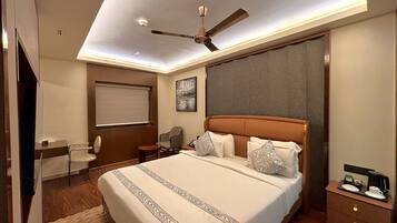 A hotel room with a large bed, a ceiling fan, a desk with a chair, a window with a blind, and a painting on the wall.