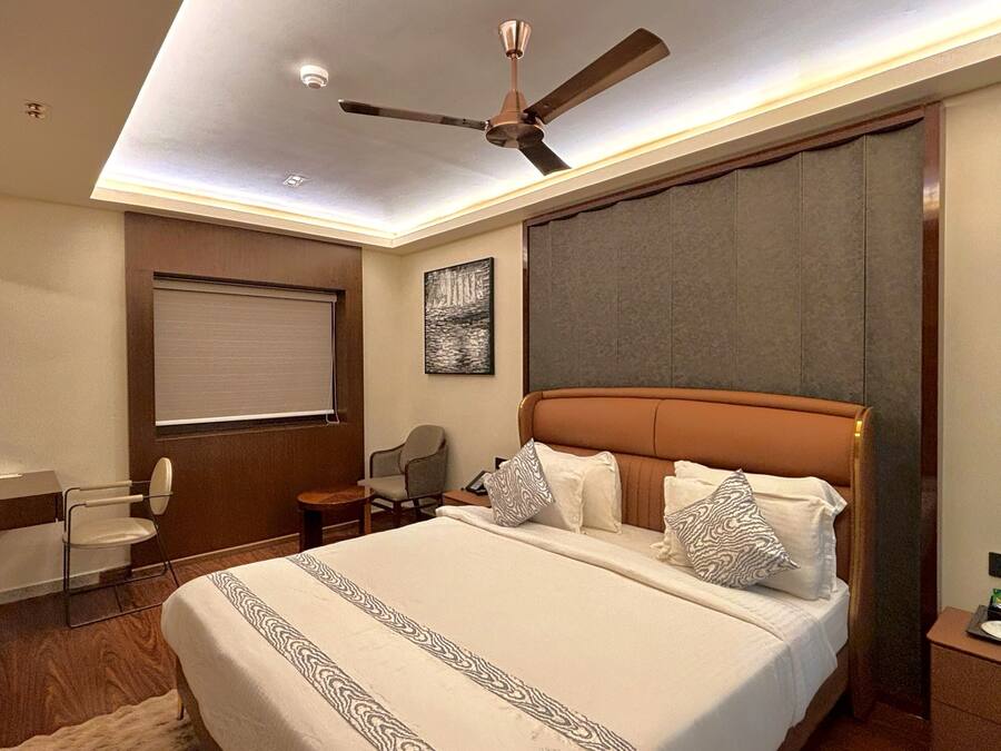 A hotel room with a large bed, a ceiling fan, a desk with a chair, a window with a blind, and a painting on the wall.